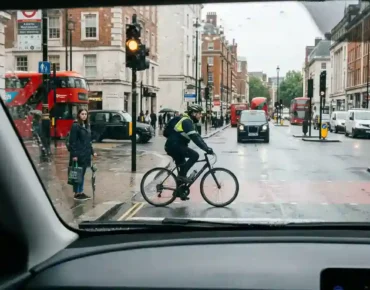 Learner driver approaching a cyclist and pedestrian at a rainy London crossing, illustrating hazard perception and driver awareness